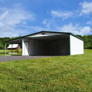 A 42X40X14/11 Vertical Roof Barn with a 12' wide lean-to, featuring a large empty white metal carport and green roof, stands in a grassy field under a blue sky with clouds. Trees and a distant building form the background.