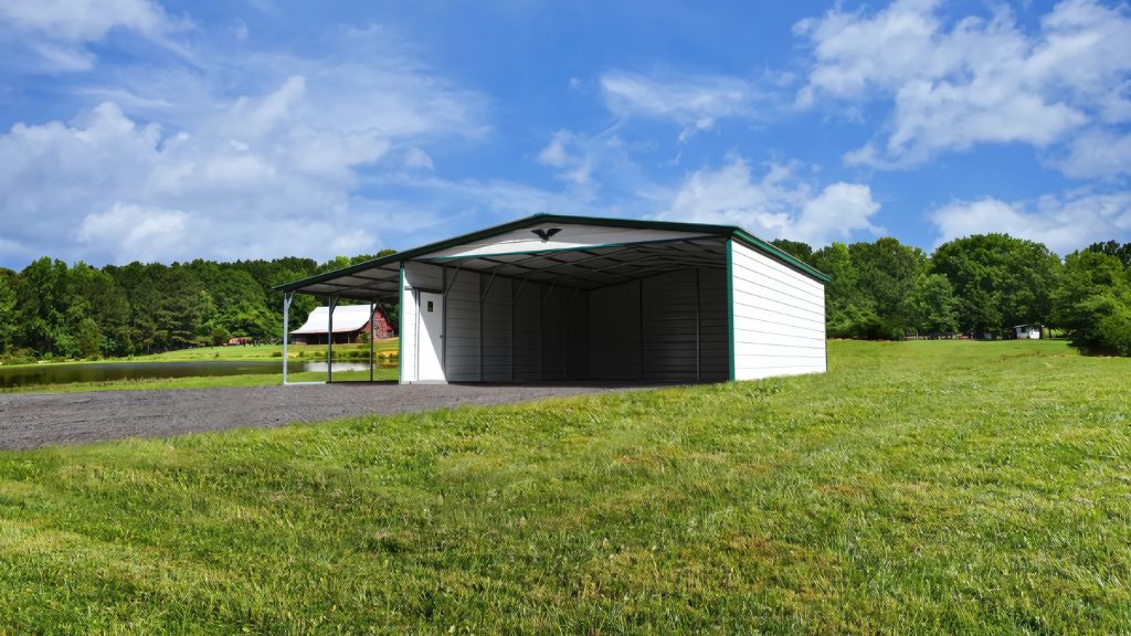 A 42X40X14/11 Vertical Roof Barn with a 12' wide lean-to, featuring a large empty white metal carport and green roof, stands in a grassy field under a blue sky with clouds. Trees and a distant building form the background.