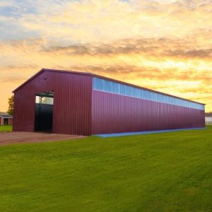 A large red barn with a vertical roof stands on a lush green field under a cloudy sunset sky, next to a white fence and an additional 40x60x12 Vertical Roof Commercial Metal Building in the background.