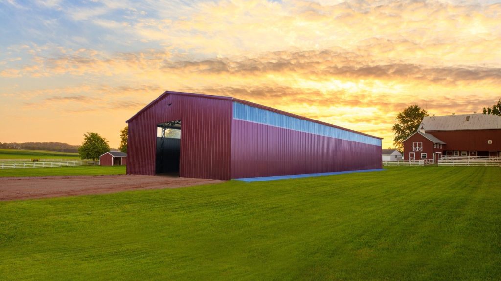A large red barn with a vertical roof stands on a lush green field under a cloudy sunset sky, next to a white fence and an additional 40x60x12 Vertical Roof Commercial Metal Building in the background.