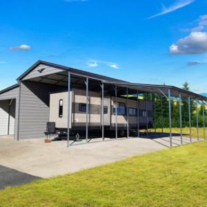 A 42X40X14/11 Vertical Roof Barn in grey metal, with a 12' wide Lean-To for an RV, rests on a concrete pad amidst trees and distant hills under a blue sky.