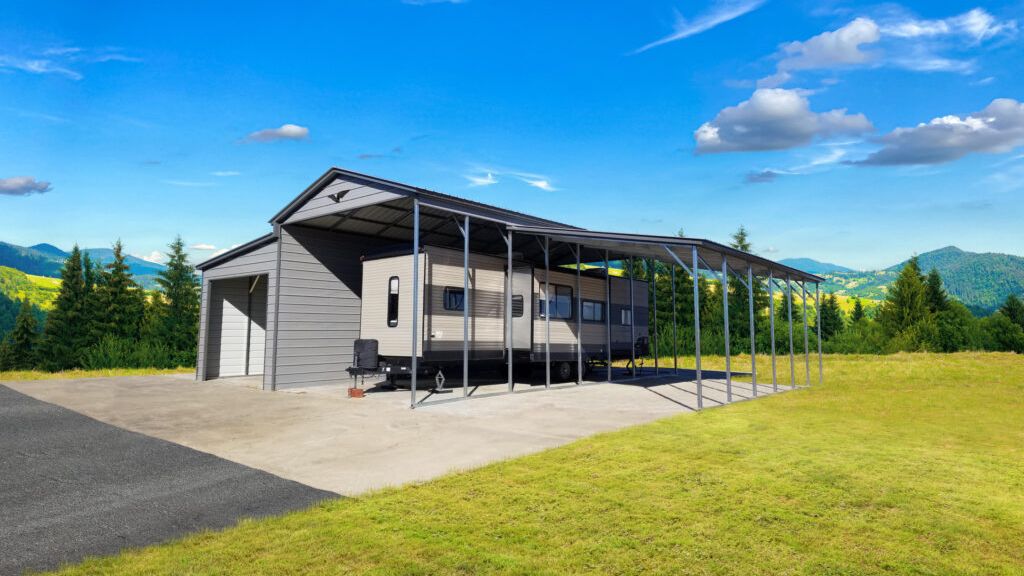 A 42X40X14/11 Vertical Roof Barn in grey metal, with a 12' wide Lean-To for an RV, rests on a concrete pad amidst trees and distant hills under a blue sky.